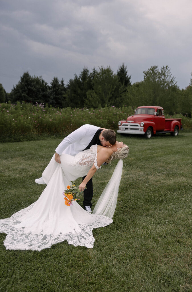 Groom dipping the bride for a romantic kiss in front of a vintage red truck at the 1885 Barn in Ohio, captured by Cleveland-based wedding photographer.