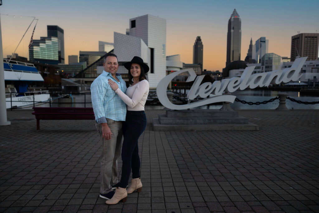 Engaged couple posing in front of the Cleveland script sign at Voinovich Park with the lakefront in the background.