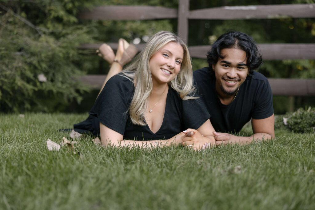 Smiling couple lying on the grass near a wooden bridge at Grand Pacific Junction in Olmsted Falls during their engagement session.