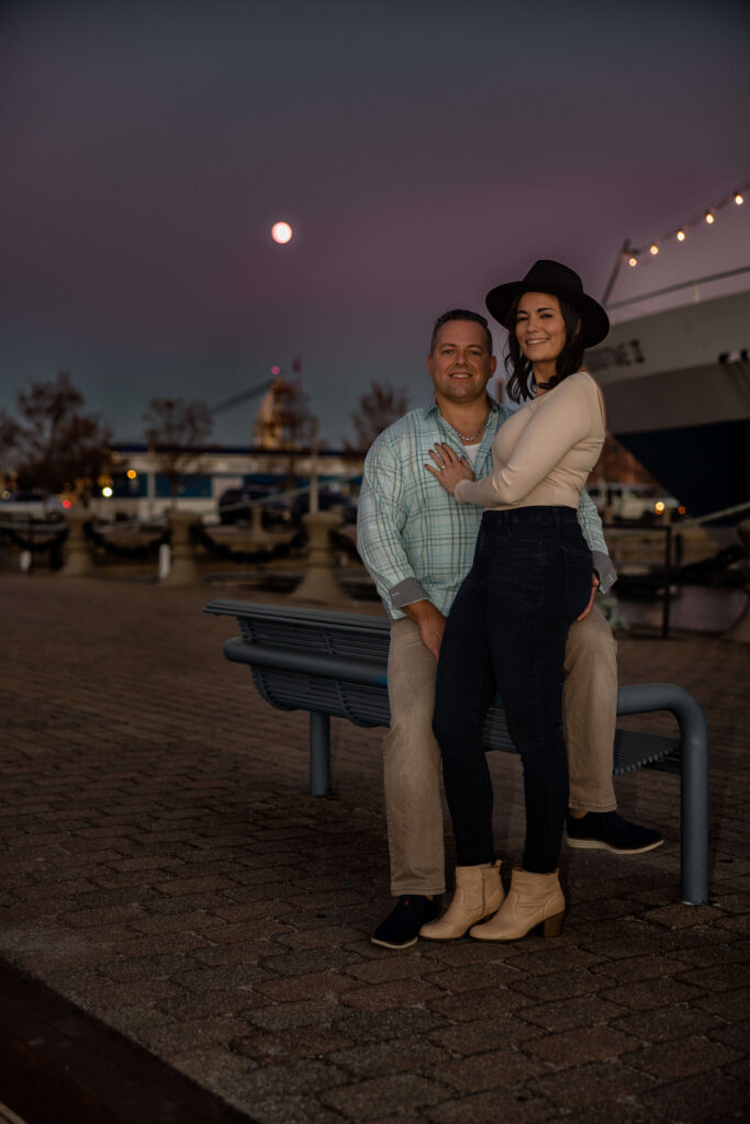 Romantic sunset engagement photo of a couple sitting on a bench at Voinovich Park with the moon and boats in the background.