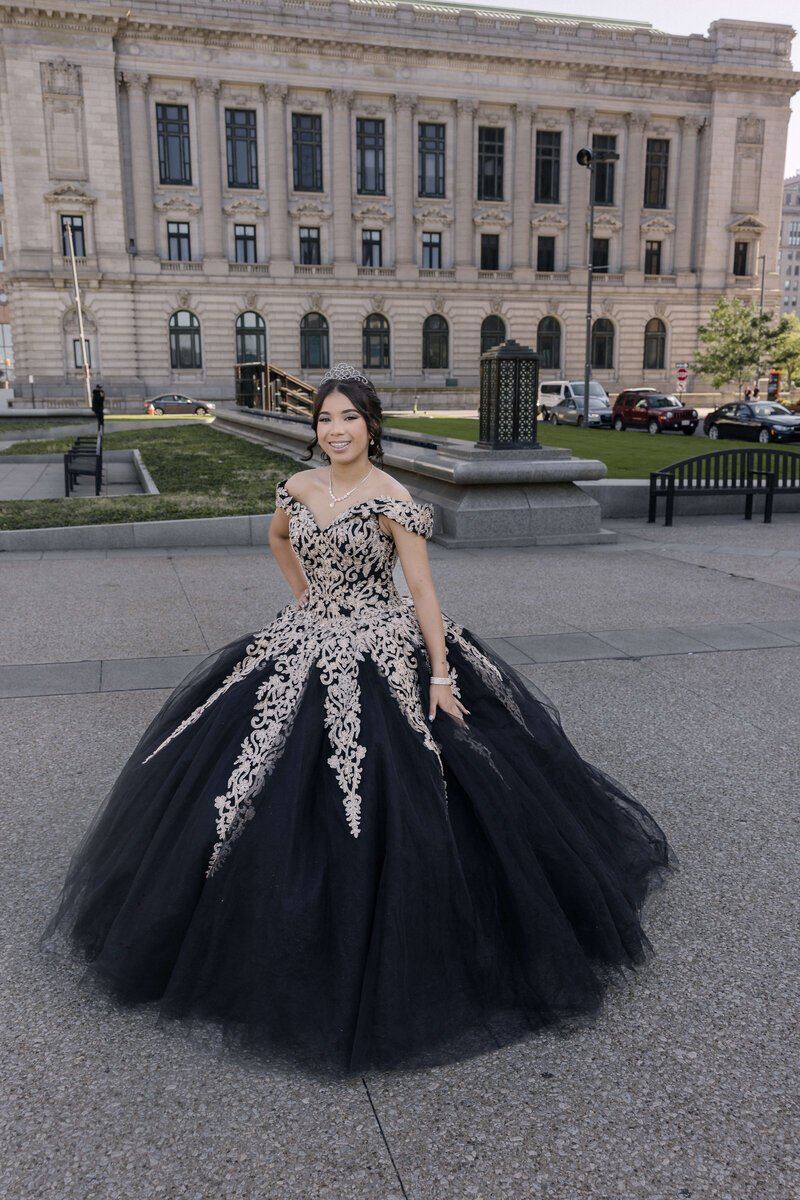 A smiling quinceañera wearing a black ball gown with champagne embroidery in front of a classical building