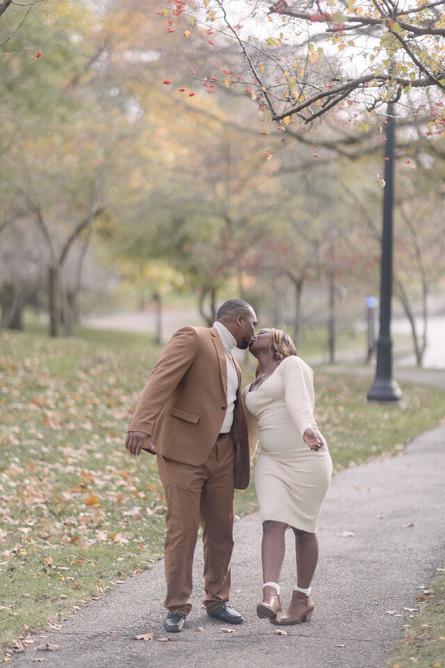 Tanisha and Kevin sharing a kiss on a park path surrounded by autumn leaves, wearing cream and camel.