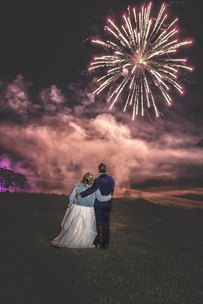 A newly married couple embracing beneath a sky filled with pink and gold fireworks